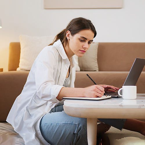 : An individual sitting on the floor, writing notes with a coffee cup and an open laptop beside them.