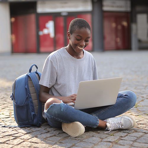 A student sitting cross-legged on the ground, looking at a laptop in their lap with a cheerful smile. A large backpack is leaning against them.