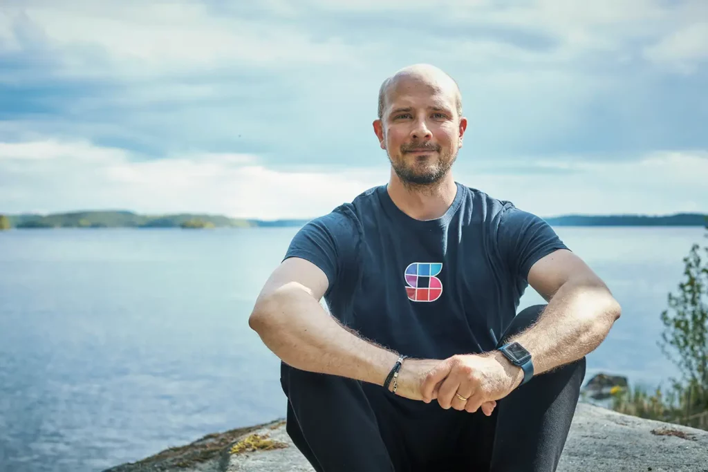 Simo Ahava sits serenely on a coastal rock, a stunning natural body of water providing a picturesque backdrop. He graces the camera with a warm smile.